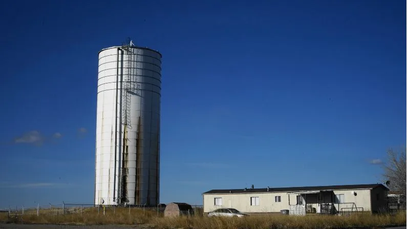 A gigantic white water tank towers above a mobile home, shed and car. They are set against the backdrop of a clear blue sky.