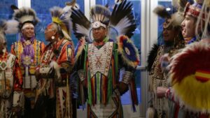 A small group of Native men dressed in traditional regalia with ornate beadwork, ribbons and feathers chat as they stand in front of tall glass windows.